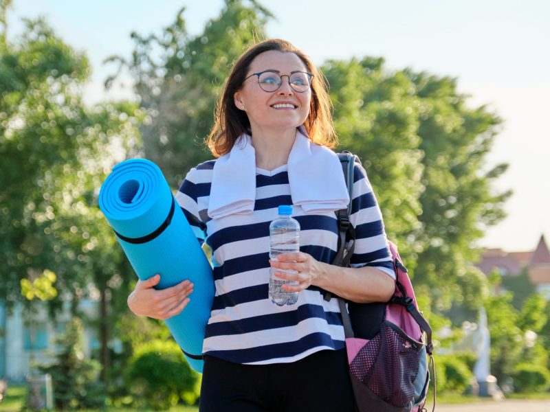 Outdoor happy mature woman walking after fit exercises in the park, with backpack, yoga mat. Active sports healthy lifestyle of middle-aged people, body and beauty